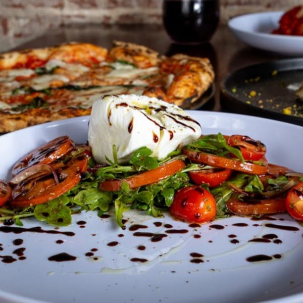 A fresh burrata salad with tomatoes, arugula, and balsamic glaze, with a pizza in the background.