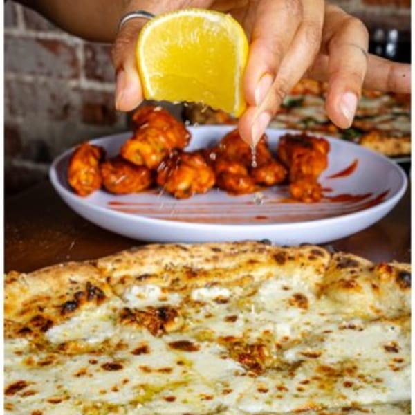 A hand squeezes a lemon over a plate of buffalo wings, with a cheesy pizza in the foreground.