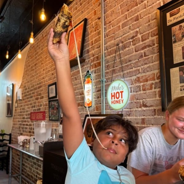 A child stretches a long string of cheese from a slice of Pie95 Pizza, with a brick wall background.