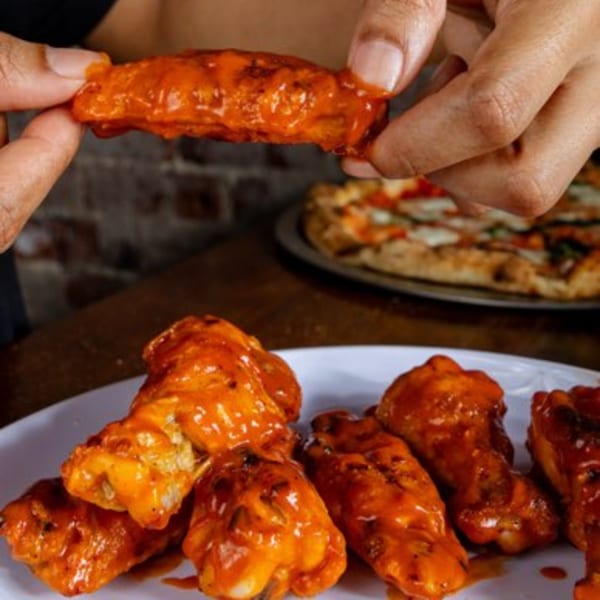 A hand holds a buffalo wing over a plate of wings, with a pizza in the background.