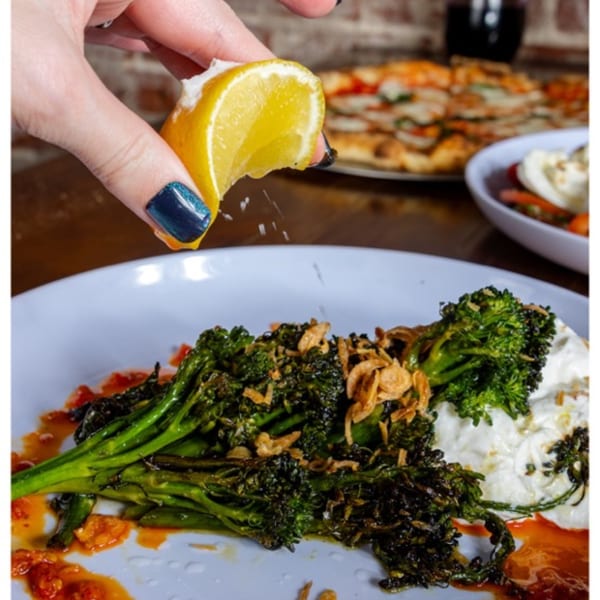 A hand squeezes a lemon over a plate of broccolini with creamy sauce and crispy onions.