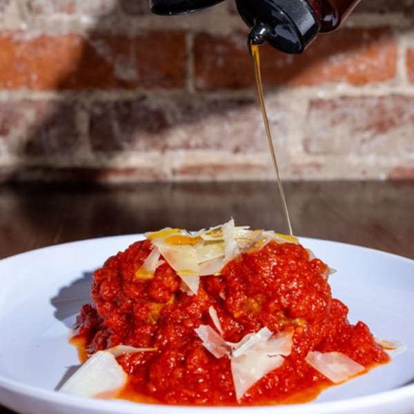 Meatballs in red sauce with shaved cheese, drizzled with honey, against a brick wall.
