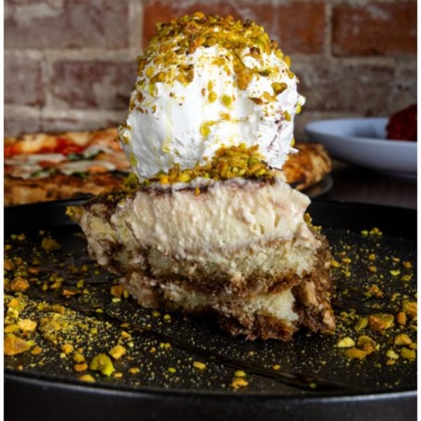 A slice of tiramisu topped with whipped cream and crushed pistachios on a dark plate.