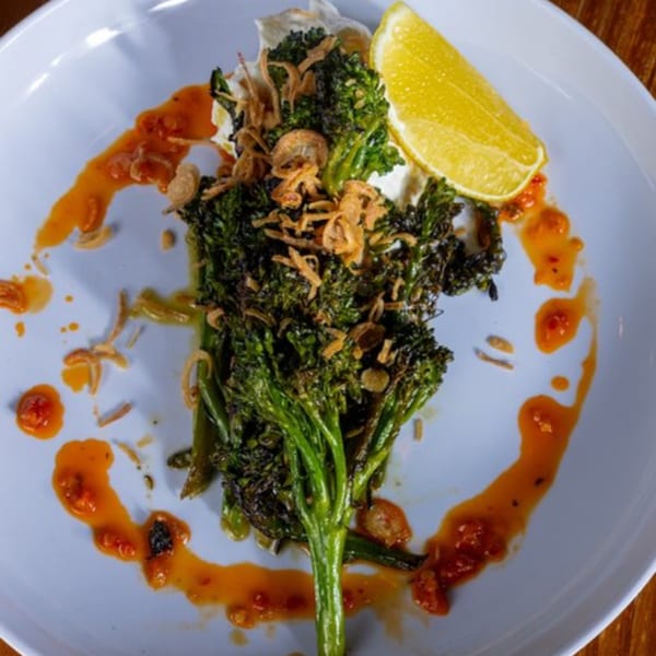Broccolini with crispy fried onions, chili oil, and a lemon wedge on a light blue plate.