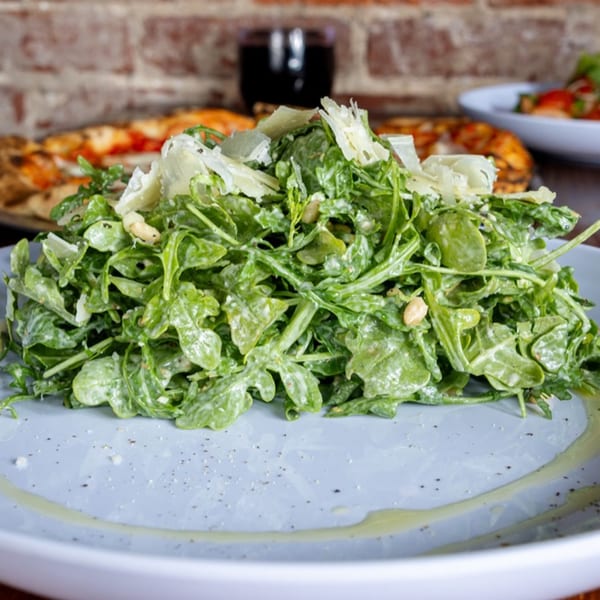 A fresh arugula salad with parmesan cheese and pine nuts, with pizzas in the background.