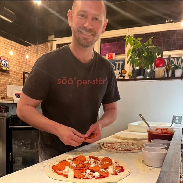A smiling man in a black t-shirt making a pepperoni pizza at Pie95 Pizza.